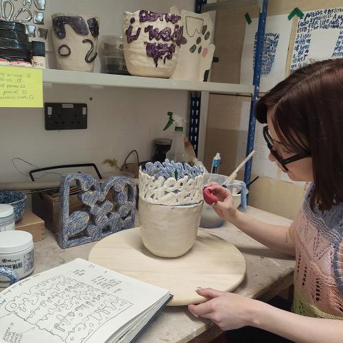 Person with shoulder-length hair and glasses scribing pottery in a studio