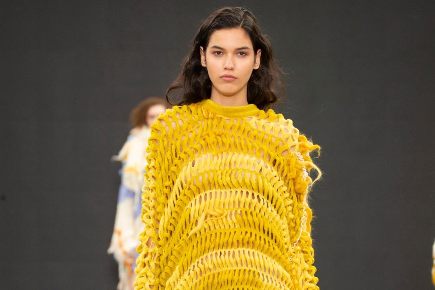 A model walks on a catwalk wearing a long yellow knitted dress