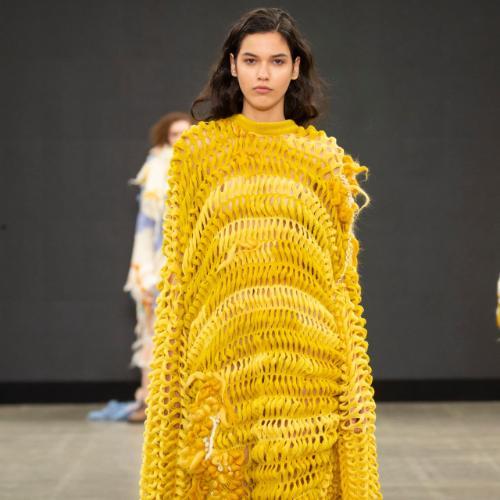 A model walks on a catwalk wearing a long yellow knitted dress