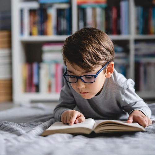 A child wearing glasses lying on a bed and reading a book, using his finger to track the words