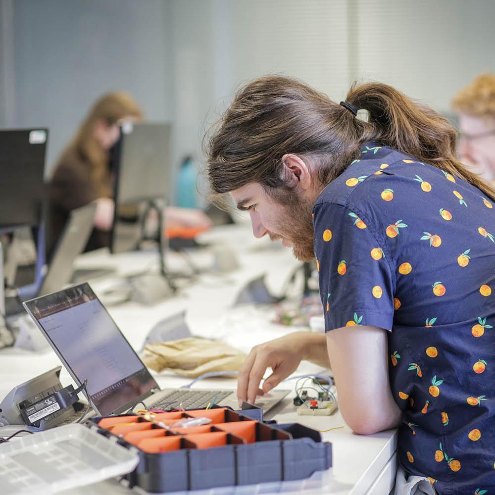 A student wearing a blue shirt with orange fruits on it working at a laptop.