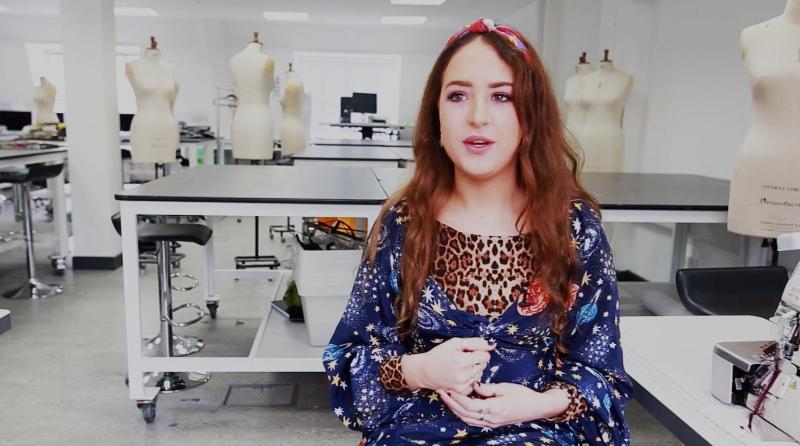 Student sitting in sewing studio, with tailors dummies and machines behind her
