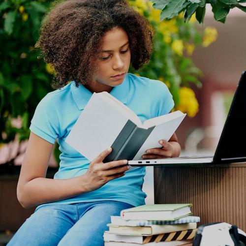 A girl sits outside reading a book with a laptop placed in front of her