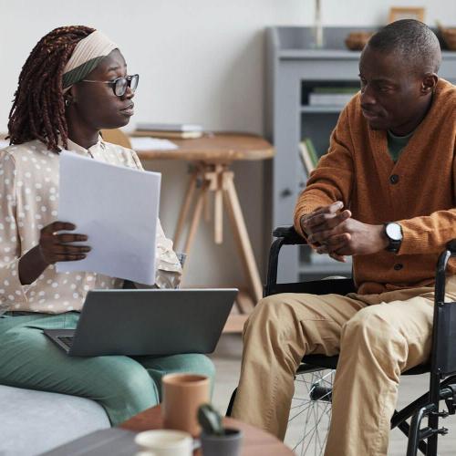 Woman with a laptop and paperwork sits next to and talks to a man in a wheelchair