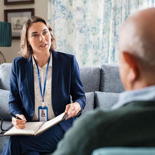 Woman in a blazer takes notes while talking to an elderly man in his home