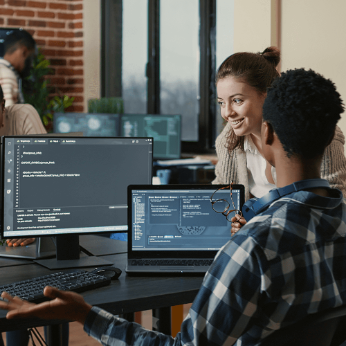 Two coworkers look at three computer screens of coding language and discuss