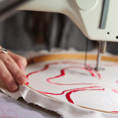 A student using a sewing machine to embroider a red freehand design onto a piece of cloth within a hoop