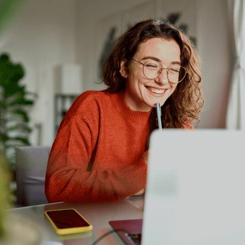 Happy young woman using laptop sitting at desk writing notes whilst looking at laptop screen learning online.