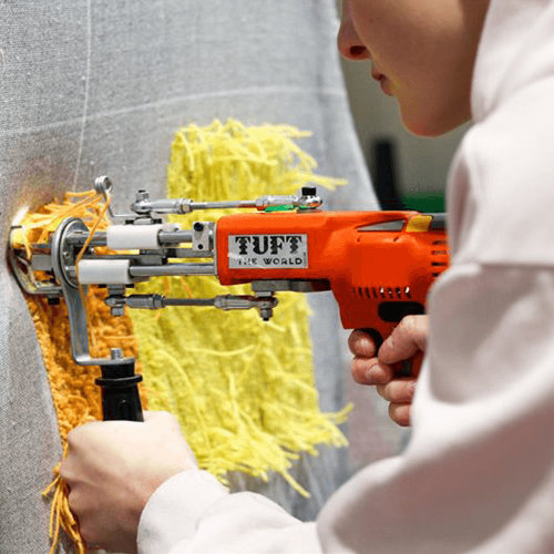 A student using an electric tufting gun to create a yellow and orange design on a piece of fabric.