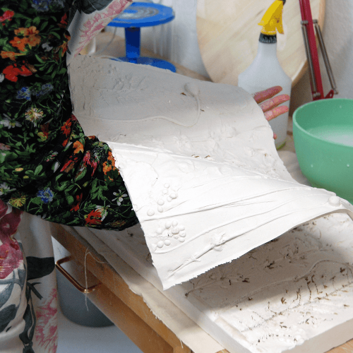 A person holds a sheet of clay with sculpted plants inlaid on top