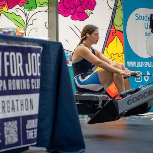 student in blue and navy rowing kit rows on a static rowing machine in the background. The foreground shows a banner reading 'Row for Joe' 