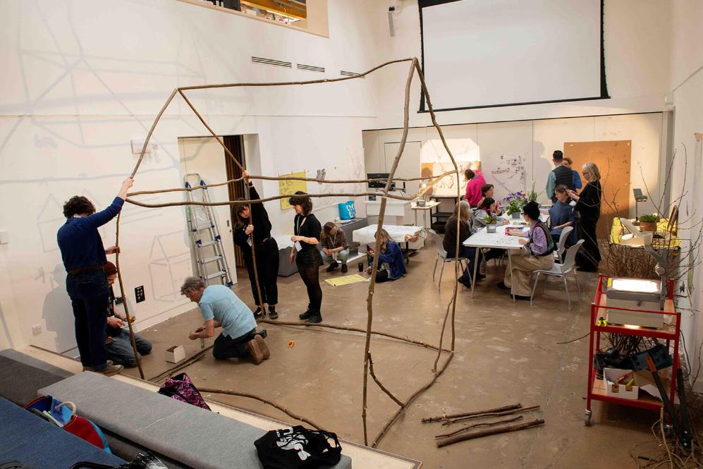 people focus on building a house frame out of wood, more gather around a table in the background.