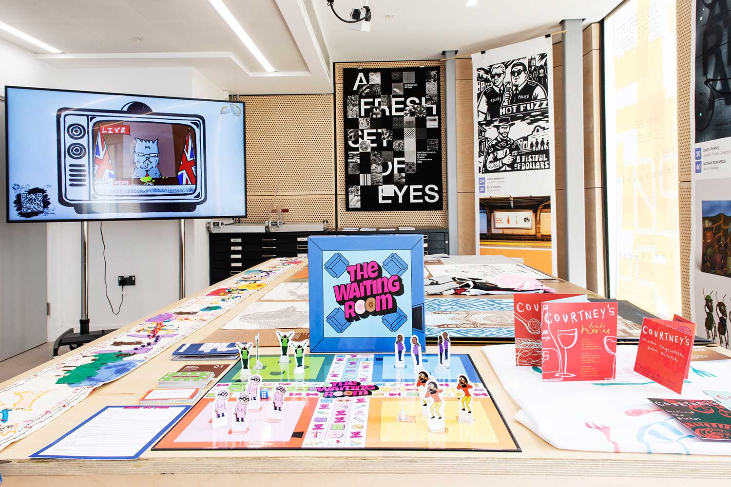 An exhibition space with graphic design work laid on tables, pinned to boards and on a digital screen