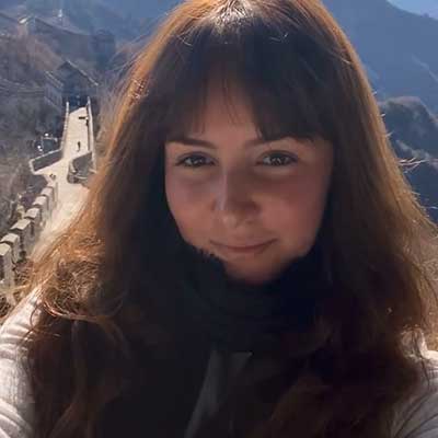 A person with long brown hair takes a selfie in front of a mountain range