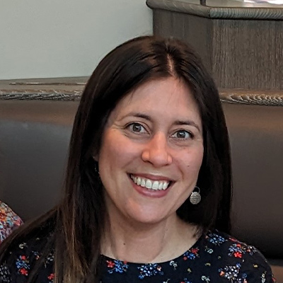 Headshot of someone with long dark hair smiling. Wearing a black floral top