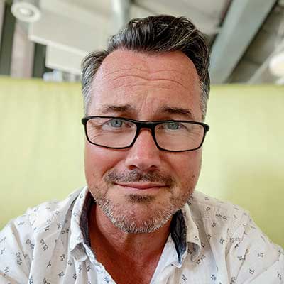 A person white short hair and black glasses looks at the camera
