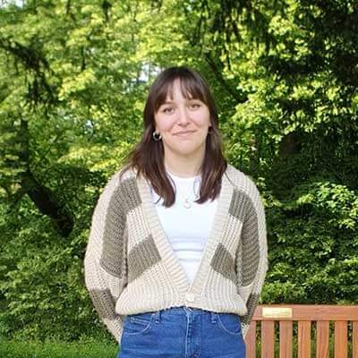A student stands in front of a green backdrop of trees and smiles at the camera